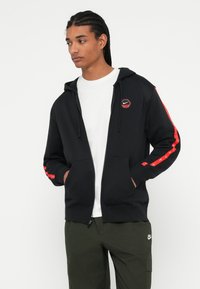 Férfi fonott hajjal, fekete Nike cipzáras kapucnis pulóverben piros csíkokkal és foltokkal, fehér pólóban és olajzöld Nike nadrágban, kezei a zsebében.