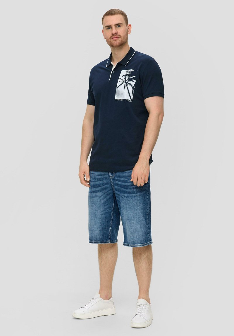 Granatowa koszulka polo z grafiką palmy na lewej piersi, w połączeniu z dżinsowymi szortami i białymi trampkami. Casualowy, luźny krój.
