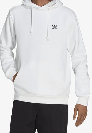 Uomo che indossa una felpa bianca Adidas con tasca anteriore e coulisse, abbinata a pantaloni neri, in piedi davanti a uno sfondo bianco.