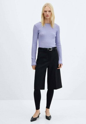Helles lila, geripptes Langarmoberteil, kombiniert mit schwarzen Shorts bis zum Knie und schwarzen Leggings. Ein schwarzer Gürtel und spitze Schuhe runden das Outfit ab.