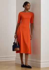 Sieviete, tērpusies pieguļošā oranžā midi kleitā ar pusgarajām piedurknēm, tur rokās tumši zilu un brūnu somu, stāvot pie baltām iekštelpu sienām.