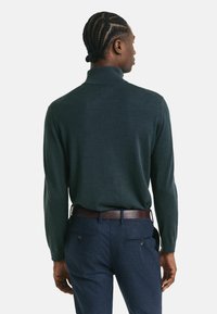 Mann mit geflochtenem Haar, der einen dunkelgrünen Rollkragenpullover und navyblaue Hosen mit einem braunen Gürtel trägt, steht mit dem Rücken zur Kamera.