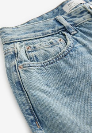 Světle modré džíny z denimu s vybledlým texturem, přední kapsou, měděnými nýty a prošitou páskovou etiketou. Zdůrazněné jemným opotřebením.