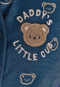 Marineblå stof med broderet design, der viser en bamse og tekst, der står "Far's lille unge" i hvid og tan tråd.