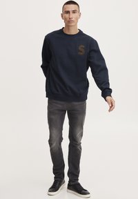 Sweatshirt bleumarin cu un logo "S" maroniu, guler rotund, mâneci lungi, asortat cu blugi gri slim-fit și adidași negri. Design simplu, textura țesăturii vizibilă.