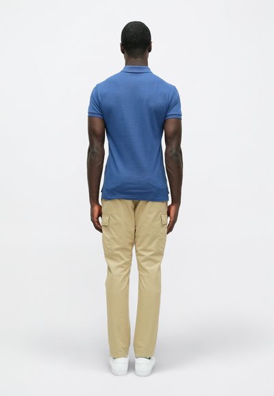 Μπλε πόλο πουκάμισο και μπεζ cargo παντελόνι, με εφαρμοστή σχεδίαση. Το πόλο έχει κλασικό γιακά και κοντά μανίκια. Λευκά αθλητικά παπούτσια.