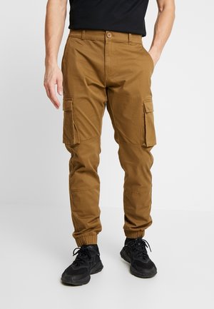 Mand iført brune cargobukser med klaplommer og elastiske manchetter, kombineret med sorte sneakers og en sort skjorte, står foran en hvid baggrund.
