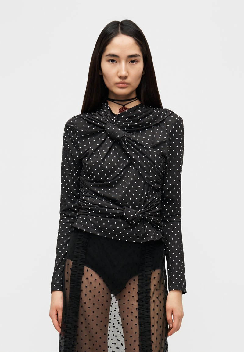 Μαύρο μακρυμάνικο τοπ με λευκές βούλες, με ριγμένα υφασμάτινα στοιχεία και απαλή υφή. Φοριέται πάνω από μια διαφανή φούστα με βούλες.