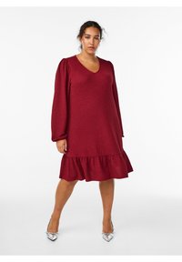 Burgundas krāsas austs kleitas ar garām pūkainām piedurknēm, V-veida kakla izgriezumu un platām viļņotām apakšmalām. Sudraba augstpapēžu kurpes papildina apģērbu.