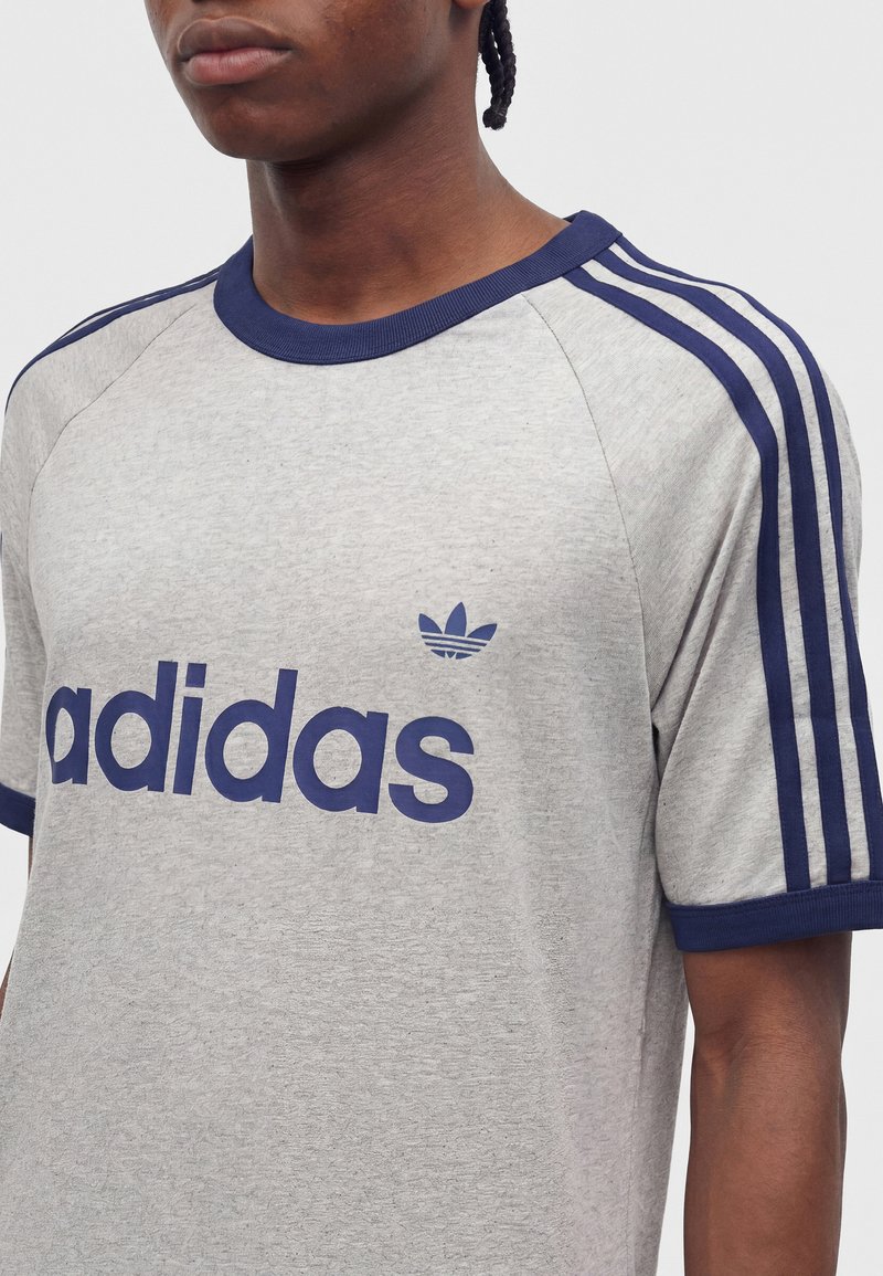 Person trägt ein graues Adidas T-Shirt mit marineblauem Kragen, Ärmelabschluss, drei Streifen auf den Schultern und Adidas-Logo auf der Brust.