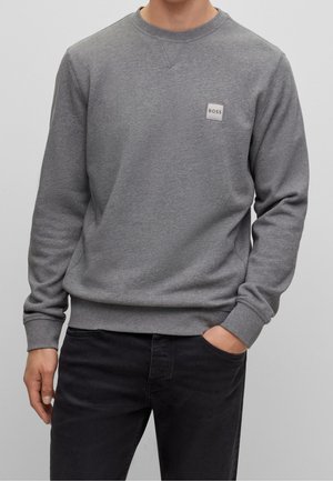 Man wearing a plain grey long-sleeve sweatshirt with a small "BOSS" logo on the chest and black jeans with one hand in pocket.