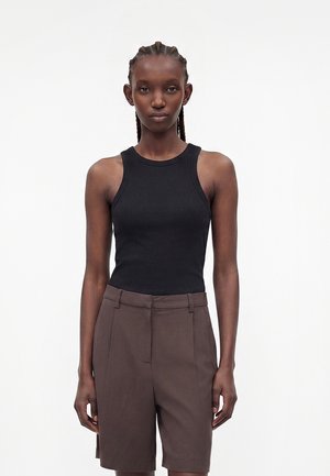 Žena s vrkočmi, oblečená v čiernom bezrukávovom top a hnedých kraťasoch s vysokým pásom, stojaca pred jednoduchým bielym pozadím.