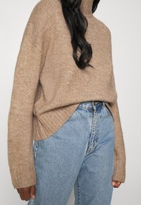 Pull à col roulé en tricot texturé marron avec des détails côtelés, associé à un jean en denim bleu clair taille haute avec une coupe classique.