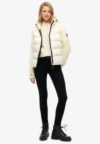 Biała kurtka puchowa z zapięciem na suwak i czarnymi akcentami, noszona na kremowym swetrze w warkocze, w połączeniu z czarnymi dopasowanymi dżinsami i czarnymi butami.