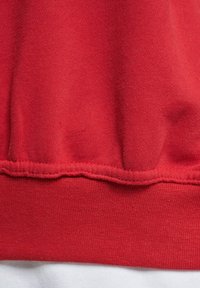 Roter Sweatstoff mit strukturiertem Oberflächen. Verfügt über einen umschlagenen Saum, der eine nahtlose Verarbeitung und leichte Raffung zeigt. Glatte weiße Fütterung sichtbar.