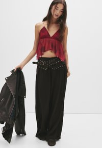 Donna che indossa un crop top senza maniche rosso e pantaloni larghi neri con una cintura borchiata, mentre tiene una giacca di pelle nera nella mano sinistra.