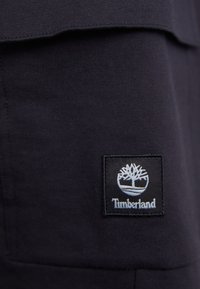 Melna kokvilnas auduma ar Timberland logotipa uzlīmi ar koka dizainu. Tuvplāns, kas parāda tekstūru un šuvju detaļas.