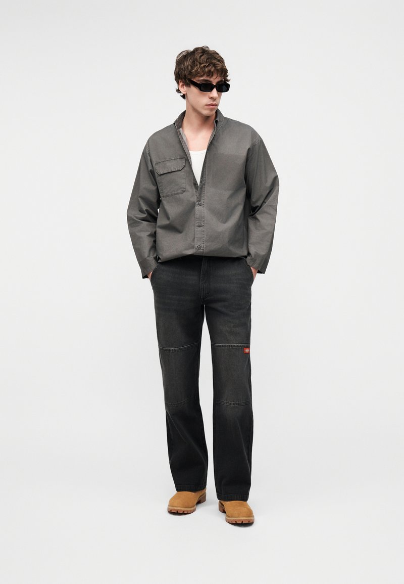 Giovane uomo con occhiali da sole neri, camicia grigia abbottonata sopra una maglietta bianca, pantaloni neri cuciti e stivali beige, in piedi con le mani nelle tasche.