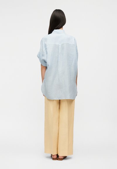 Femme aux longs cheveux noirs portant une chemise ample à rayures bleues et blanches et un pantalon large beige, debout de dos face à la caméra.