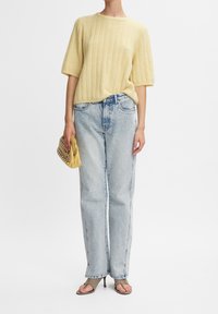 Person som bär en ljusgul kortärmad stickad topp, högmidjade ljusblå raka jeans, håller en gul kuvertväska och bär taupe öppna klackskor.