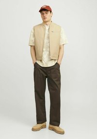 Beige ärmellose Weste über einem hellen gemusterten Hemd, kombiniert mit dunkelbraunen Hosen und hellbraunen Stiefeln, ergänzt durch eine braune Mütze.