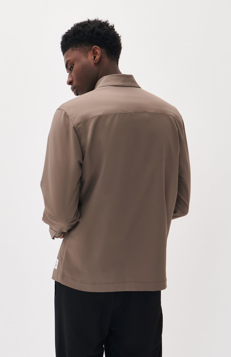 Veste marron légère à coupe décontractée, dotée d'un col, de manches retroussées et d'un design simple. Tissu texturé et étiquette discrète visibles.