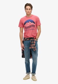 Camiseta roja jaspeada con gráfico de montaña, jeans de mezclilla, cinturón negro y camisa de franela a cuadros atada a la cintura. Zapatillas de deporte beige.