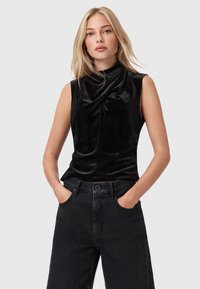 Blonde vrouw in een mouwloze zwarte fluwelen top met hoge hals en zwarte high-waisted wijde broek, handen in de zakken, neutrale achtergrond.