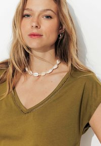Haut vert olive en V à manches courtes avec des détails en dentelle le long de l'encolure ; accessoirisé avec un collier en coquillage blanc.