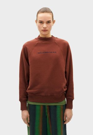 Felpa marrone con colletto alto e maniche raglan. Testo "ECCO ARRIVARE IL SOLE" in blu marino, tessuto morbido, polsini e orlo a costine.