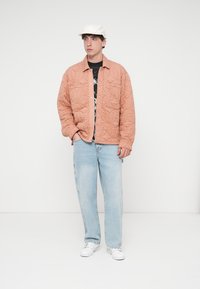 Ραμμένο ροδακινί μπουφάν με κουμπιά στο μπροστινό μέρος, φορεμένο πάνω από μαύρο t-shirt με σχέδιο, συνδυασμένο με ανοιχτόχρωμο τζιν και λευκά αθλητικά παπούτσια. Συμπληρώνεται με λευκό καπέλο.