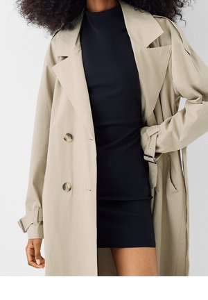 Femme portant un trench-coat beige sur une robe noire ajustée, une main sur la hanche et les cheveux bouclés visibles.