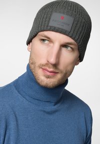 Szara, prążkowana czapka beanie z prostokątną czarną nalepką z czerwonym logo. Noszona z niebieskim golfem. Gładka tekstura, dopasowany krój.