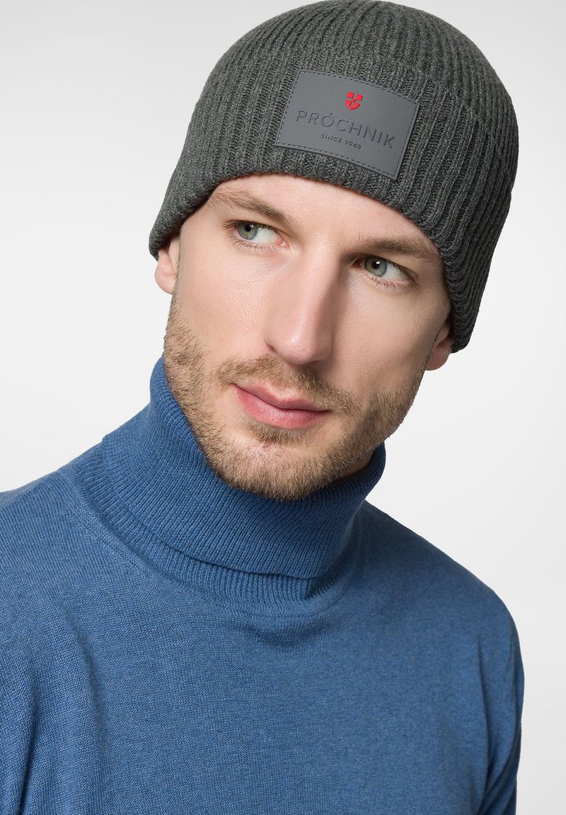 Szara, prążkowana czapka beanie z prostokątną czarną nalepką z czerwonym logo. Noszona z niebieskim golfem. Gładka tekstura, dopasowany krój.