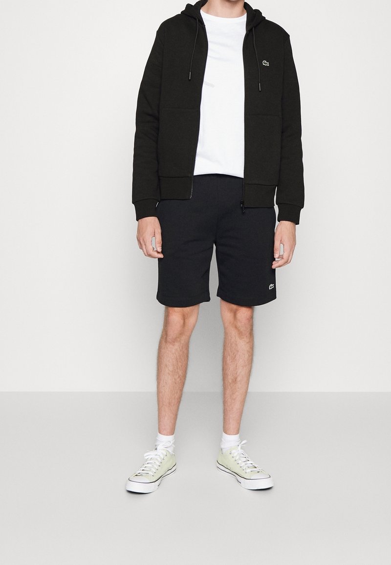 Homme portant un sweat à capuche noir zippé, un t-shirt blanc, un short noir, des chaussettes blanches et des baskets claires sur un fond uni.
