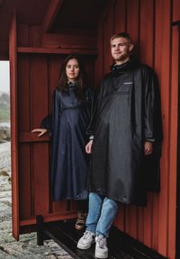 Två individer med vattentäta ponchos, en marinblå och en svart, står mot en röd träbakgrund. Ponchosarna har huvor och oversize-passform.