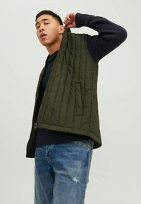 Grüne gesteppte Weste mit Reißverschluss und Seitentaschen, über einem marineblauen Pullover getragen. Das Material wirkt leicht und strukturiert.