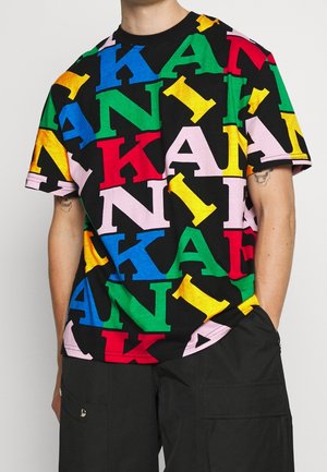 Mies, joka käyttää mustaa t-paitaa, jossa on suuria, värikkäitä kirjaimia "K", "A", "N" ja "I" sinisen, vihreän, punaisen, vaaleanpunaisen ja keltaisen sävyissä, päällekkäin.
