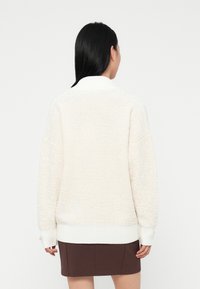 Person with long black hair wearing a white textured sweater with ribbed cuffs and hem, and a dark brown skirt, facing away on a white background.
