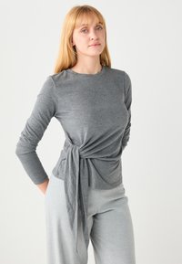 Haut gris à manches longues avec un détail noué à la taille, confectionné dans un tissu doux, associé à un pantalon gris clair pour un look harmonieux.