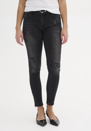 Mujer con jeans pitillo negros desgastados y dobladillos con cremallera, una blusa blanca, un cinturón negro y zapatos slingback negros de punta afilada.