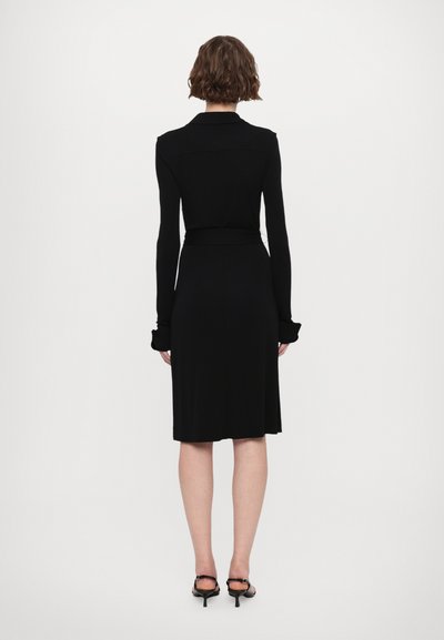 Čierne šaty s dlhým rukávom a priliehavým siluetom, s golierom a opásaným pásom. Hladká textúra a dizajn do výšky kolien, doplnené čiernymi podpätkami.