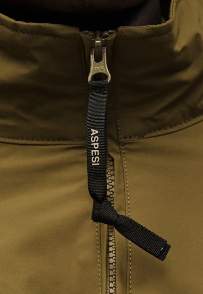 Közeli kép egy olívazöld kabát cipzárjáról, fekete szövet húzófüllel, amin az "ASPESI" felirat látható, és fém cipzár csúszkával, melyen a "YKK" jelzés szerepel.