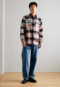 Camicia di flanella a quadri nera, arancione e azzurro chiaro con due tasche sul petto. Jeans blu a vestibilità ampia e scarpe nere completano l'outfit.