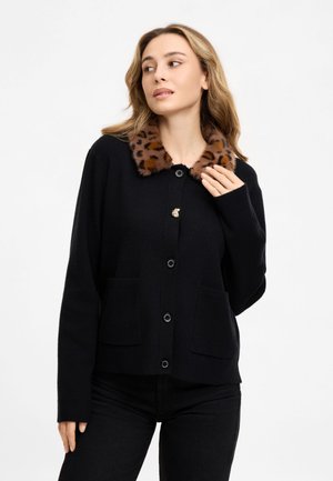 Frau trägt eine schwarze, zum Knöpfen geschnittene Strickjacke mit einem braunen Leopardenmuster und Kunstpelzkragen, stehend vor einem schlichten weißen Hintergrund.