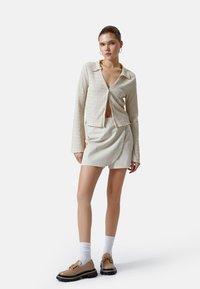 Camicia lungo beige chiaro e texturata con colletto, abbinata a una gonna crema e scarpe marroni con suola nera spessa.