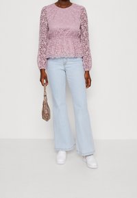 Lila spetsöverdel med långa, genomskinliga ärmar och midja med volang, kombinerad med ljusblå utsvängda jeans och vita sneakers. Håller en ormskinnsväska.