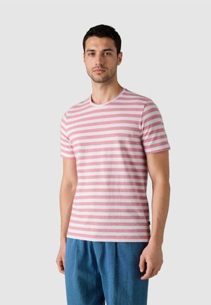 Hombre vestido con una camisa de manga corta a rayas horizontales rosa y blanca, y pantalones azules, de pie frente a un fondo gris claro.