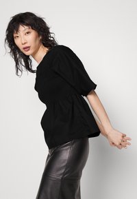 Top preto com mangas bufantes e corpete franzido, apresenta uma bainha godê fluida. Combinado com uma saia justa de couro preto, textura suave, design minimalista.