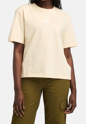 Kobieta ubrana w kremowy t-shirt z krótkim rękawem i oliwkowe spodnie typu cargo, stoi z ramionami opuszczonymi wzdłuż boków.